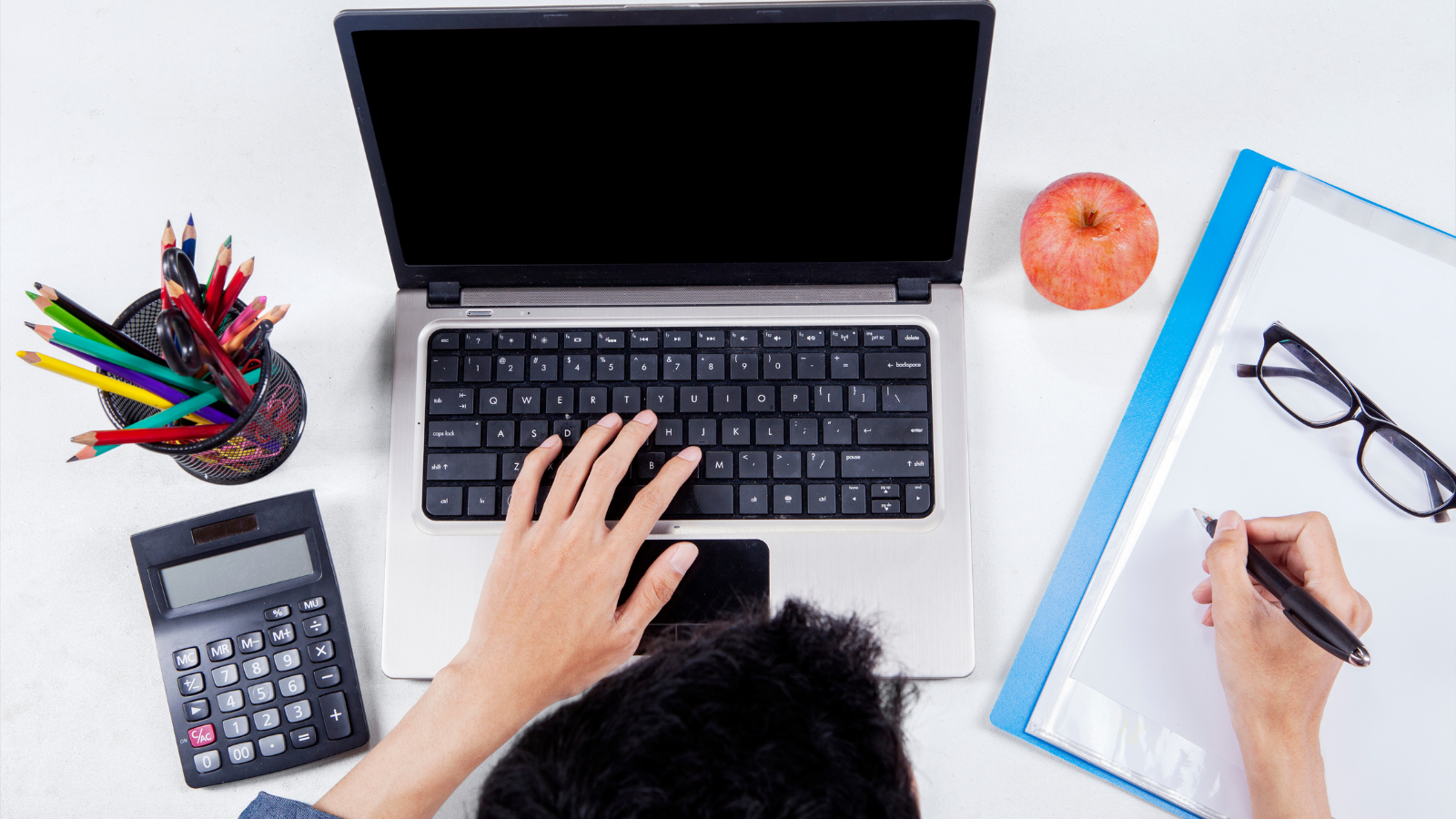 Person working on a table with laptop, papers, calculator, pens, apple and glasses